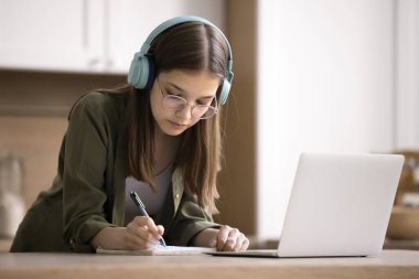 Odaklanmış genç kız evde sınava hazırlanıyor, mutfakta masada duruyor, kablosuz kulaklıklar üzerine sesli dersler dinliyor, notlar yazıyor, internetten yabancı dil öğreniyor, internet üzerinden ders çalışıyor.