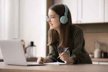 Düşünceli genç kız ödev yapıyor, kablosuz kulaklık takıyor, evde ders çalışıyor, bilgisayardan ses kurslarını dinliyor, başka yere bakıyor, kompozisyon fikrini düşünüyor, not alıyor, yeni bilgiler kazanıyor, öğrenme sürecinin tadını çıkarıyor.