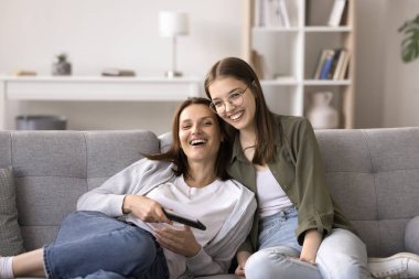 Neşeli anne ve genç kız kanepede rahatlayın, gülümseyin, iletişim kurun, uzaktan kumandayla vakit geçirin, akıllı televizyonda kanal değiştirin, favori komediyi izleyin, modern teknolojinin ve tembel hafta sonunun tadını çıkarın.