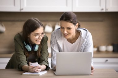 Akıllı telefonu elinde tutan ve olgun bir anneyle konuşan güzel bir genç kız okul ödevine dizüstü bilgisayarla yardım ediyor, mutfakta birlikte ayakta durmaya hazırlanıyor. Modern teknolojiyi ve uygulamaları kullanarak eğitim