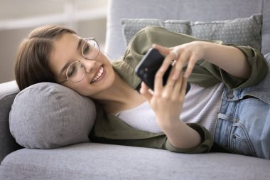 Güzel genç kız kanepede telefonla uzanıyor, arkadaşlarıyla sohbet ediyor, internette geziniyor, mesaj yazıyor, modern aletlerle vakit geçiriyor, yeni uygulamalar öğreniyor, sosyal ağda video seyrediyor, kapatıyor.
