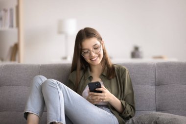 Gülümseyen ergen kız akıllı telefonuyla rahatlar, kanepede oturur, oturma odasında tek başına vakit geçirir, arkadaşlarıyla sohbet eder, mesaj yazar, mesaj okur, sosyal ağlara bakar, modern teknoloji kullanımından zevk alır.