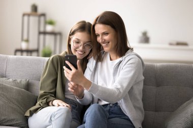 Mutlu genç kız rahat bir kanepede olgun annesiyle rahatlar, birlikte zaman geçirir cep telefonu kullanır, interneti gezer, yeni uygulamalarla eğlenir, sıcak ilişkilerin tadını çıkarır, sosyal ağda video izleriz.