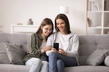 Gülümseyen genç kız annesiyle vakit geçiriyor rahat bir kanepeye oturuyor, cep telefonuyla eğleniyor, yeni uygulama kullanıyor, sosyal ağlarda video izliyor, internetten ilginç haberler okuyor, bilgi arıyor.