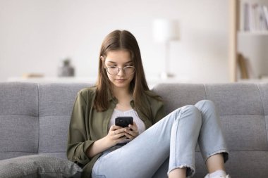 Genç kız akıllı telefonuyla kanepede oturur, mesajlaşır, arar, arkadaşlarıyla ve aileleriyle iletişimde kalmak için mesajlaşma uygulamaları kullanır, güncellemeleri paylaşır, fotoğraf ve videoları paylaşır, platformda diğerleriyle iletişim kurar.