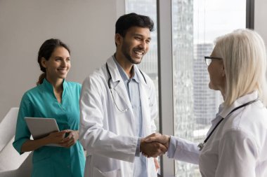 Genç erkek stajyer, beyaz önlük giyip orta yaşlı kadın doktorla el sıkışıyor, birlikte duruyor, karşılıyor, birbirimizi gördüğümüze seviniyor, klinikte başarılı bir takım çalışmasıyla tebrik ediyor.