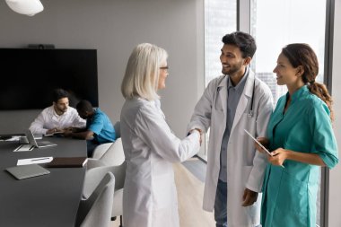 Beyaz önlüklü deneyimli bayan doktor, Hintli genç erkek doktorla el sıkışıyor, hoş geldin jestleri yapıyor veya anlaşma yapıyor, profesyonel ortaklığa başlıyor, akıl hocalığı yapıyor, veya başarılı tartışmalar yapıyor.