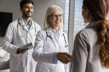 Olgun kadın doktor ve stajyer klinik hastaları selamlıyor, el sıkışıyor, misafirperverlik gösteriyor, konsültasyona başlıyor, kliniği bitiriyor, profesyonel tıbbi bakım ve yardım veriyor. Sağlık, iletişim.