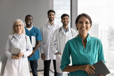 Klinikte meslektaşlarıyla poz veren, kameraya bakıp gülümseyen güzel İspanyol kadın dahiliyeci takım çalışmasından, iş birliğinden, kariyer büyümesinden ve görevden memnun. GP portreleri