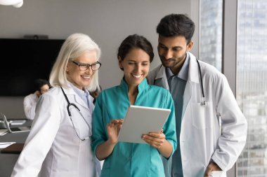 Tablet kullanan, işbirlikçi tartışma ve başarılı takım çalışması yapan, çevrim içi çalışan üç gülümseyen doktor, cihaza odaklanan içeriğe odaklanan, yeni modern teletıp uygulaması öğreniyorlar.