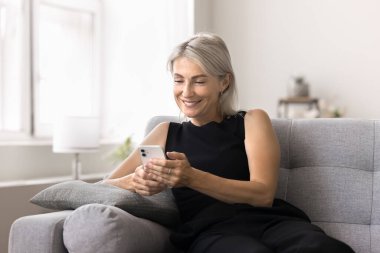 Olgun bir kadın evdeki koltukta oturan akıllı telefonu kullanır, görevleri yönetir, etkinlikleri ayarlar, online alışverişin keyfini çıkarır, resimleri düzenler veya yeni mobil uygulama kullanarak sosyal medya için içerik oluşturur.