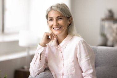 Portrait of attractive middle-aged female in casual pink blouse smile look away resting sits on comfortable couch in living room, spend carefree leisure at home, deep in pleasant thoughts and memories clipart