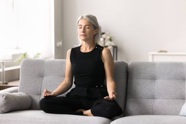 Olgun, huzurlu bir kadın dinleniyor, evde meditasyon yapıyor, koltuğa oturmuş stresi azaltıyor, farkındalığı ve farkındalığı arttırıyor, duygusal refahı arttırıyor, ruhsal gelişimi ve bağlantıyı geliştirmek için bir yöntem buluyor.