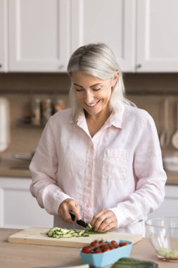 Orta yaşlı neşeli bir kadın diyet salatası için taze salatalık doğruyor, aile için mutfakta sağlıklı bir akşam yemeği hazırlıyor, yemek pişirme sürecinden zevk alıyor, dengeli beslenmeye devam ediyor. Ev yapımı yemekler, vejetaryenlik.