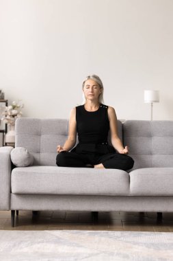 Nilüfer pozisyonu almış, kanepeye oturmuş meditasyon yapan sakin bir kadın. Sakin zihni geliştirmek ve vücudu rahatlatmak, stresi azaltmak, genel refahı teşvik etmek, odaklanmayı ve konsantrasyonu geliştirmek için pratik yapın.