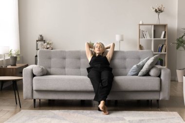 Huzurlu, orta yaşlı, çekici bir kadın rahat bir kanepeye uzanmış, uykulu görünüyor, şık bir oturma odasında tek başına şekerleme yapıyor. Rahatlama, yorgunluk nöbeti, hafta sonları sağlıklı dinlenme.