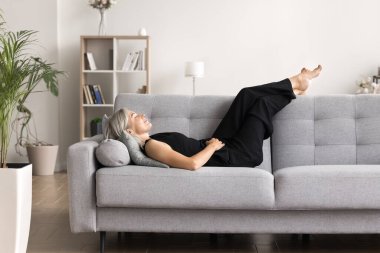 Olgun bir kadın uzun bir günün ardından dinlenmek ve dinlenmek için zaman ayırıyor, modern oturma odasındaki rahat kanepeye uzanıyor, enerjisini yeniliyor ve evde yalnız başına kaygısızca eğleniyor.