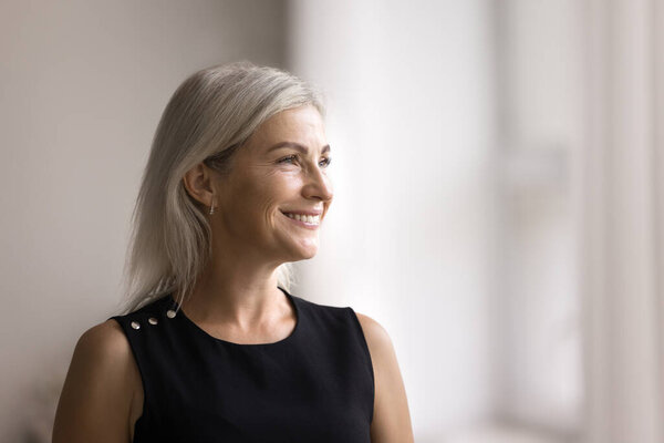 Head shot beautiful middle-aged female smiling looking out window, standing alone in modern living room, admire view from apartment, deep in pleasant thoughts, recollect, enjoy warm memories at home