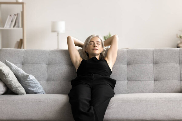 Serene middle-aged woman closes her eyes put hands behind head breath fresh conditioned air inside fashionable living room, enjoy stress-free day off alone at own or rented flat. Fatigue relief, peace