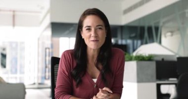 Genç 35 'lik İspanyol kadın modern ofiste masa başında oturuyor, kameraya bakıyor, video çağrı uygulaması kullanarak konuşma yapıyor, video görüşmelerine katılıyor, çevrimiçi brifing veriyor, proje güncellemelerini uzaktan yapıyor.