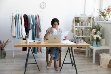 Gülümseyen Kafkasyalı genç iş kadını moda tasarımcısı modern ofis masasında oturup sabah kahve içmeyi düşünür. Mutlu kadın stilisti ya da evrak işleriyle uğraşan terzi iş yerinde dizüstü bilgisayar kullanın.