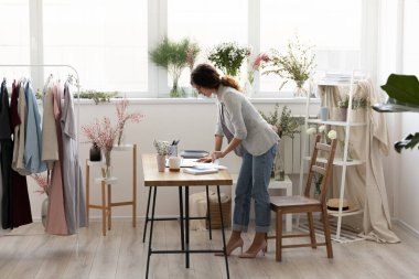 Başarılı genç beyaz kadın tasarımcı kendi modern stüdyo işyerinde çalışıyor. Avrupalı kendine güvenen kadın moda tasarımcısı veya terzisi atölye veya galeride yeni kıyafet örnekleri veya koleksiyonu hazırlar.