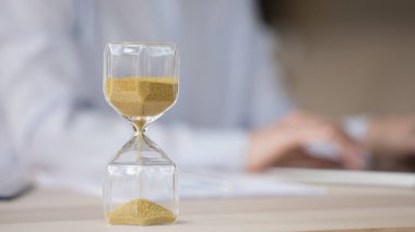 Kum saati objesi, insan eli bilgisayarın arkasında bulanık bir şekilde yazarken yakın çekim yapıyor. Çalışma masasında kum akan kum saati. İş, zaman yönetimi, son teslim tarihi kavramı
