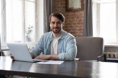 Başarılı hırslı girişimci, iş sahibi portre konsepti. Millennial erkek ofis çalışanı kameraya bakıyor çalışma masasına oturuyor, laptopta çalışıyor, modern teknoloji kullanıyor, kurumsal yazılım kullanıyor.