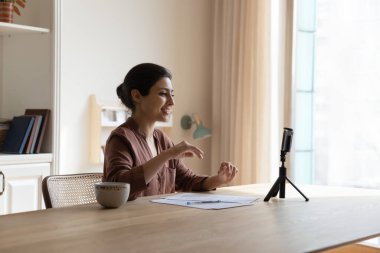Gülümseyen Hintli kadın video kaydediyor, tripoda akıllı telefon kullanıyor, ahşap masada oturuyor, mutlu genç bayan iş kadını blogcu ya da öğretmen web kamerasından canlı yayında konuşuyor, internette sohbet ediyor.