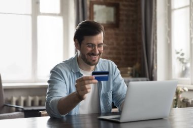 Başarılı bir e-ödeme, ani para transferi, e-alışveriş, güvenli e-banka konsepti. Gülümseyen genç adam masada oturuyor. Bilgisayarı kullanıyor. İnternetten kredi kartı alıyor.