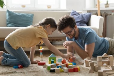 Pozitif nişanlı baba ve kız çocuğu halı kaplı zeminde küplerle oynuyor, oyuncak kule inşa ediyor, kale inşa ediyor, inşaat tuğlalarını düzenliyor, kızların hayal gücünü geliştiriyor, yaratıcılığını geliştiriyor.