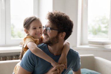 Mutlu genç baba, tatlı kızını sırtında taşıyor. Kanepede oturuyor, neşeli kızla aktif oyunlar oynuyor. Konuşuyor, gülüyor, eğleniyor, aile eğlencesinden keyif alıyor.