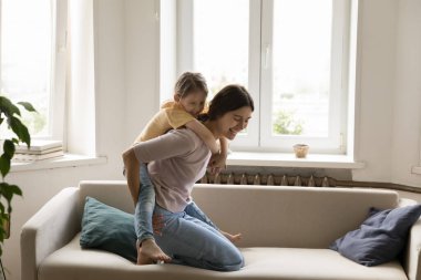 Neşeli küçük kızıyla kanepede oyun oynayan, kanepede oturan, gülümseyen ve gülen mutlu bir genç anne. Annesine binen neşeli kız