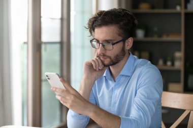 Ciddi milenyum işadamı masada oturur, cep telefonundan gelen mesajı okur, yeni iş başvurusu öğrenir, e-posta okur, modern cihazlar kullanarak internetten bilgi toplar.