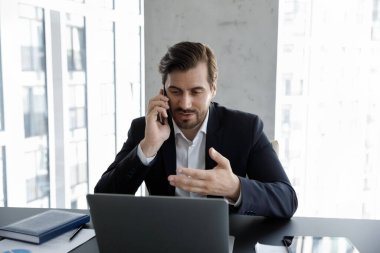 Modern gökdelen ofisinde çalışan takım elbiseli ciddi bir işadamı, masada oturan birini arıyor ve akıllı telefondan müşteriyle dizüstü konuşmalarına bakıyor. Teknoloji, iş akışı, yaşam tarzı, mesafeli iletişim kavramı