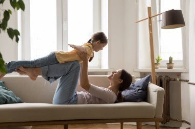 Neşeli, genç ve güçlü bir anne, küçük kızıyla uçak oyunu oynuyor, kanepeye uzanıyor, çocuğu havaya kaldırıyor, akroyoga desteği veriyor, eğleniyor, gülüyor. Yan görünüm