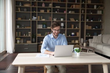 Gözlüklü genç işadamı dizüstü bilgisayarla çalışıyor, güncel oturma odasında masa başında oturuyor, mesajları paylaşıyor, e-posta yazıyor, uzaktan iş çözüyor, tele-iş yapıyor.