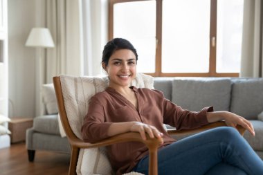 Portre gülümseyen Hintli kadın modern oturma odasındaki rahat koltukta rahatlıyor, mutlu ve çekici genç bayan ev sahibi ya da kiracı kameraya bakıyor, hafta sonları evde boş vakit geçiriyor.
