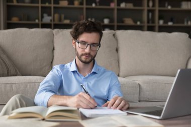 Ciddi meşgul erkek öğrenci notlar yazıyor, deneme veya özet hazırlıyor, araştırma üzerinde çalışıyor, evde oturma odasında ders kitapları ve dizüstü bilgisayarla masada oturuyor. Uzaktan öğrenme, kendi kendini eğitme