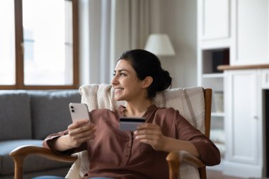 Sırıtkan Hintli kadın elinde akıllı telefon ve kredi kartı, uzaktan bakıyor, evdeki rahat koltukta oturuyor, mutlu genç bayan online alışveriş yapıyor, sipariş veriyor, internet bankacılığı hizmetlerini tarıyor.