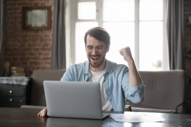 Heyecanlı milenyum serbest iş adamı iyi haberler alıyor, e-posta, dikkat, proje başarısını kutluyor, dizüstü bilgisayarda mutlu bir gülümsemeyle oturuyor, kazanan eli yapıyor, gülüyor, bağırıyor.