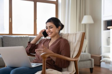 Mutlu Hintli kadın telefonda konuşuyor, dizüstü bilgisayar ekranına bakıyor, evdeki rahat koltukta oturuyor, genç bayan serbest çalışan müşteriye cep telefonuyla danışıyor, çevrimiçi çalışıyor.
