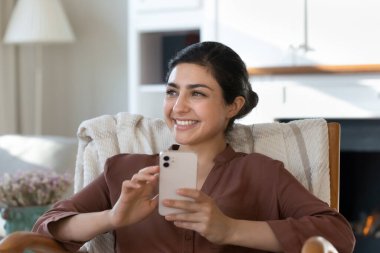 Fotoğrafta gülümseyen Hintli genç bir kadın, uzaktan bakıyor, akıllı telefondan kafası dağılmış mutlu, genç bir kadın, güzel bir geleceği hayal ediyor, başarının hayalini kuruyor, internette sohbet ediyor.