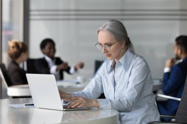 Modern iş yerinde dizüstü bilgisayar kullanan, gözlüklü ciddi orta yaşlı profesyonel kadın. Ofiste çalışan olgun bir çalışan. Arka planda toplanan iş arkadaşları