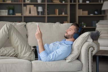 Serene 'li 30' lu yaşlardaki genç adam kablosuz modern kulaklıkla müzik dinlemekten hoşlanıyor, akıllı telefonuyla koltukta uzanıyor, pistte tek başına zaman geçiriyor ve modern teknolojiyi kullanıyor. İnternet akışı çevrimiçi hizmetler, dinlenme