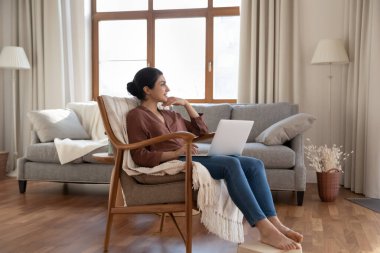 Gülümseyen rüya gibi Hintli kadın dizüstü bilgisayar kullanıyor, evinde oturuyor, uzaktan bakıyor, uzaktan bakan düşünceli genç bayan serbest çalışıyor, güzel bir geleceği hayal ediyor, başarının hayalini kuruyor.