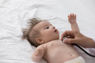 Bebek bezli güzel bir erkek ya da kız çocuğu, beyaz çarşafa uzanmış tatlı bir bebek, doktor terapistine bakıyor stetoskop kullanıyor, kalp atışlarını kontrol ediyor. Pediatri konsepti