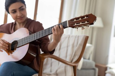 Hintli kadın akustik gitar çalıyor, akorlar çalışıyor, evdeki rahat koltukta oturuyor, genç bayan müzisyen müzik aleti çalıyor, boş zamanların tadını çıkarıyor, hobi olarak