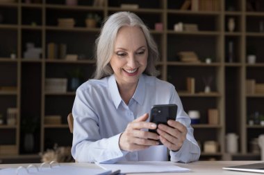 Neşeli yaşlı iş kadını evde akıllı telefon kullanıyor, çalışma masasında cihaz tutuyor, internetten sohbet ediyor, ekrana bakıyor, internetten alışveriş yapıyor. Emeklilik, kablosuz iletişim konsepti