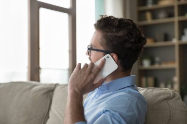 Yan görüşlü adam, akıllı telefondan konuşur, kanepede dinlenir. Gözlüklü genç adam evde yalnız başına oturup uzaktan kişisel konuşmalara bakıyor, uzaktan düzen kuruyor. Modern teknoloji, iletişim.